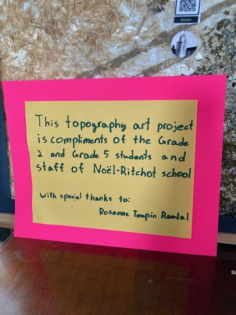 Sign reading: "This topography art project is compliments of the Grade 2 and 5 students and staff of Noël-Ritchot school. With thanks to: Rosanne Toupin Ramlal"